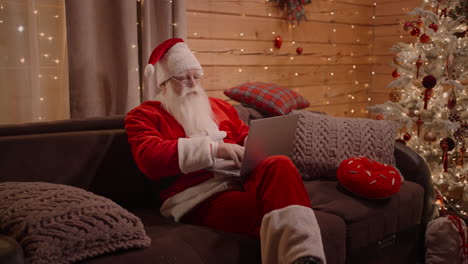 Santa-relaxes-at-home-near-a-decorated-Christmas-tree-with-a-laptop-and-answers-messages.-Santa-Claus-on-the-Eve-of-Birth-At-Home